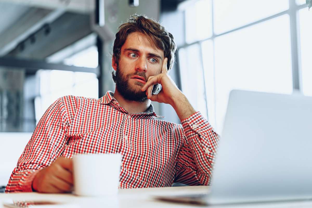 LaunchInFive-Website-Design A man with a beard and wearing a red checkered shirt sits at a desk looking at a laptop. He is holding a white mug in one hand and appears deep in thought, with his other hand resting on his face. The background shows a modern office setting.