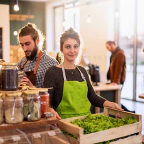 pride Two people working in a shop filled with jars of food items. The person in the foreground, wearing a green apron, holds a crate of green leafy vegetables and smiles at the camera. The person in the background, also in an apron, is focused on the jars.