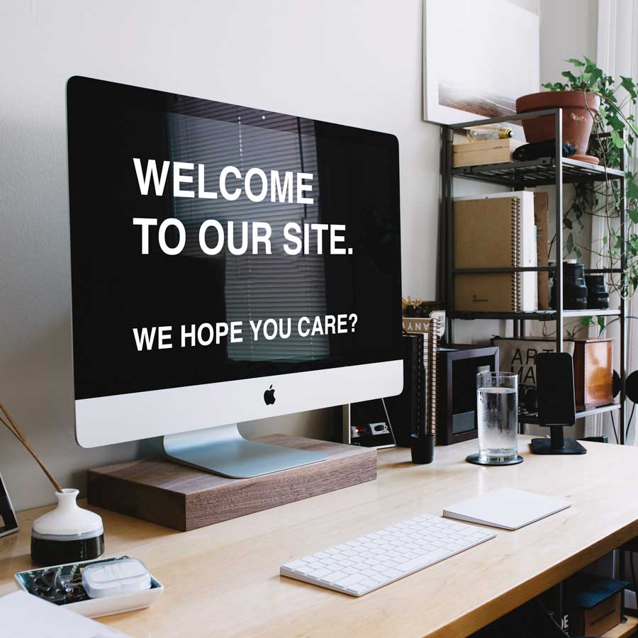 welcome A desktop computer on a wooden desk displays the text "WELCOME TO OUR SITE. WE HOPE YOU CARE?" The desk also holds a keyboard, mouse, smartphone, notebook, glasses, and some decorative plants. Shelves with books and more plants are in the background.