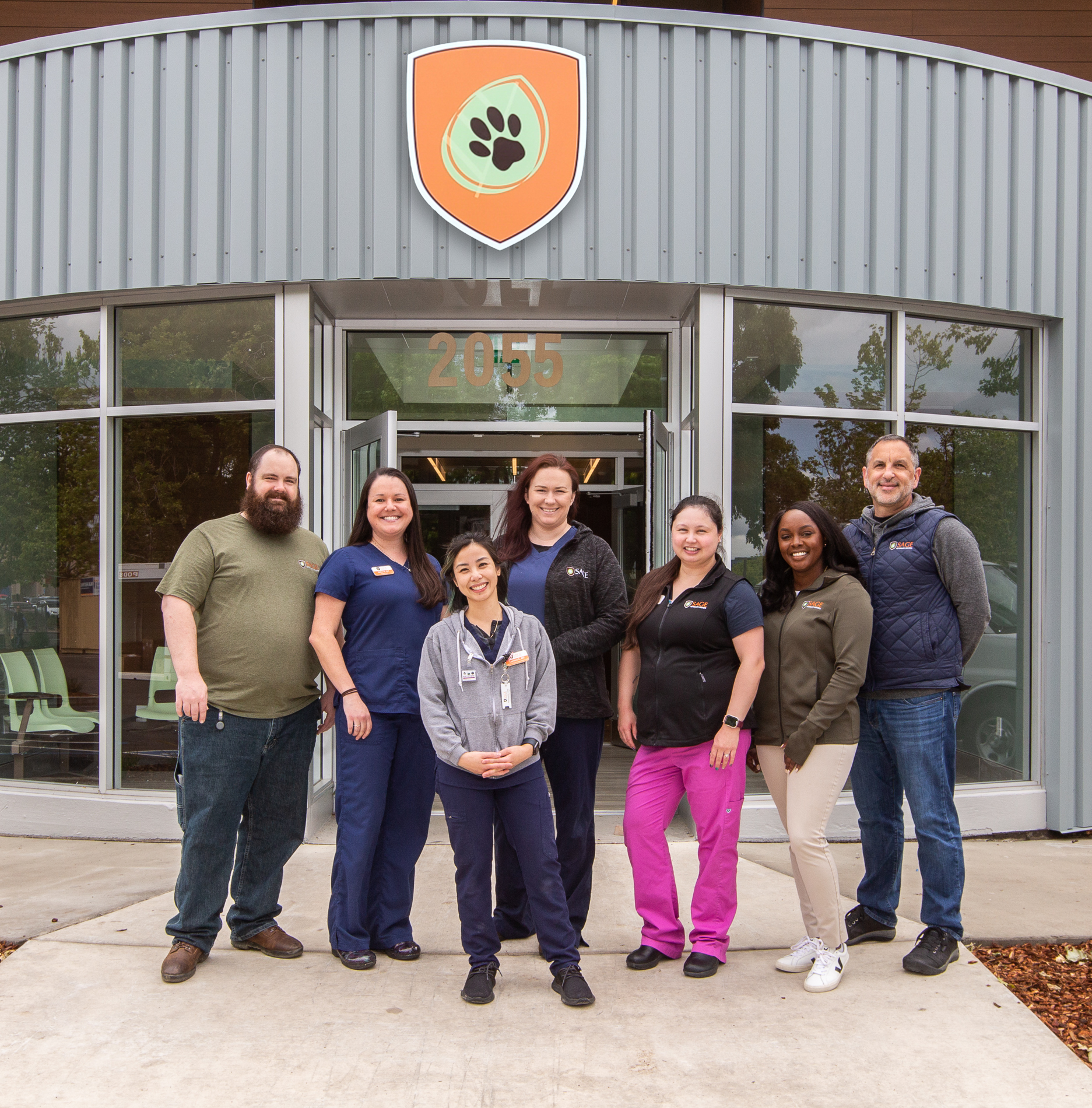 -01-5 A group of seven people, casually dressed and some in medical scrubs, stand outside the entrance of a building. The building has a large shield with a paw print above the door and the number "2055" above the entrance.