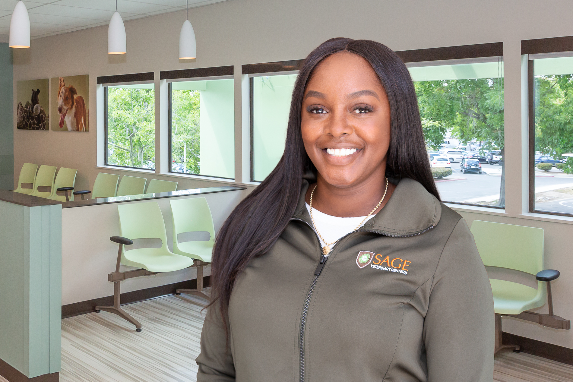 Elonia Butler A woman wearing a green jacket with an embroidered "SAGE Veterinary Centers" logo stands smiling in a modern veterinary clinic's waiting area. The clinic features large windows with views of greenery, green chairs, and pet artwork on the walls.