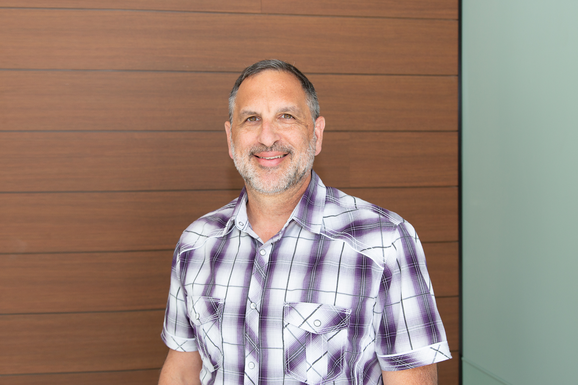 Jeremy Renshaw A middle-aged man with short gray hair and a beard is smiling at the camera. He is wearing a short-sleeved purple plaid shirt. The background features a wooden panel wall.