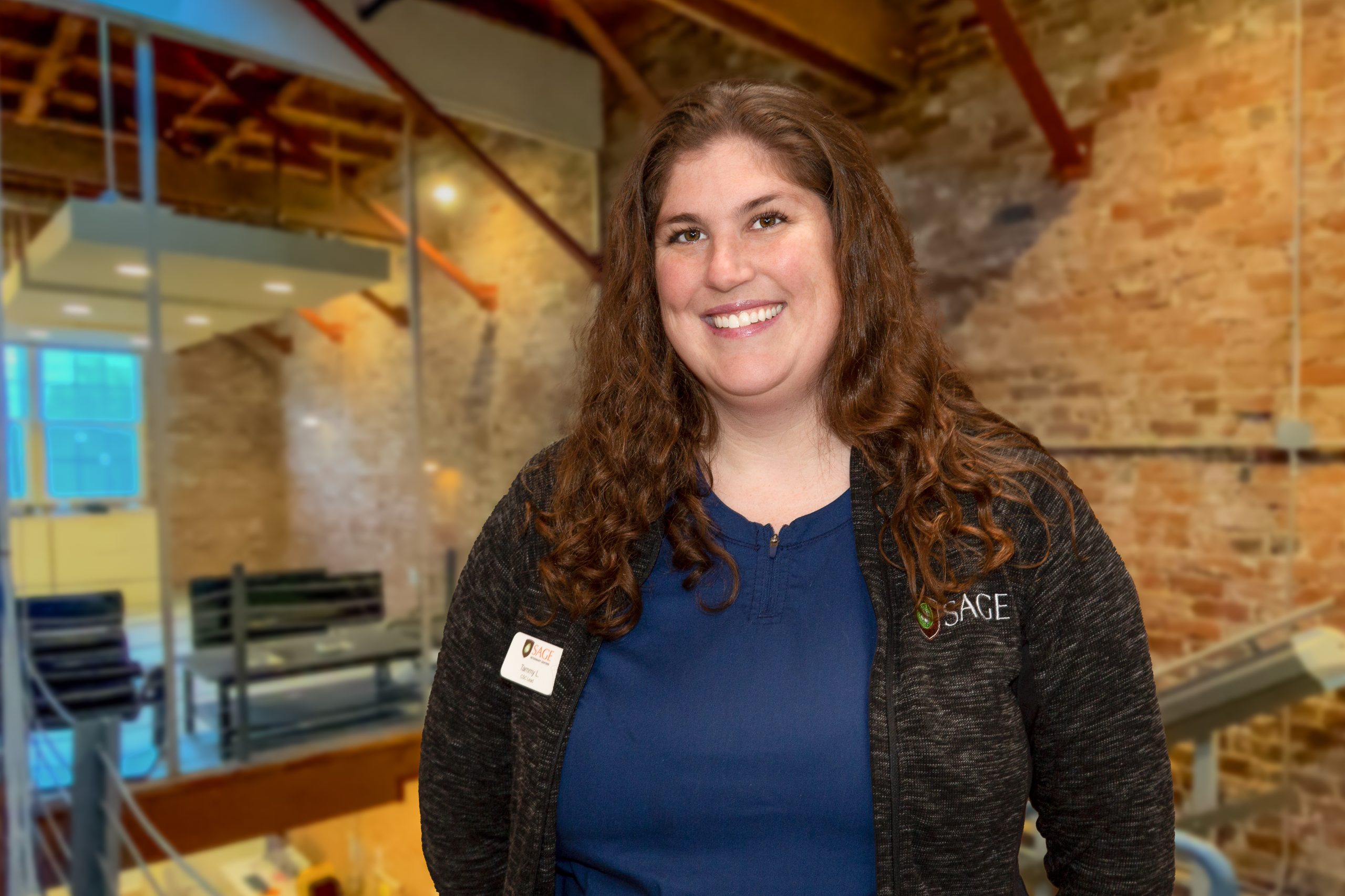 Jessica-Lorenzetti-RVT A woman with long curly hair, wearing a dark zip-up jacket over a blue top, smiles at the camera. She has a name tag on her jacket and stands in an industrial-style room with exposed brick walls and metal beams. The background is slightly blurred.