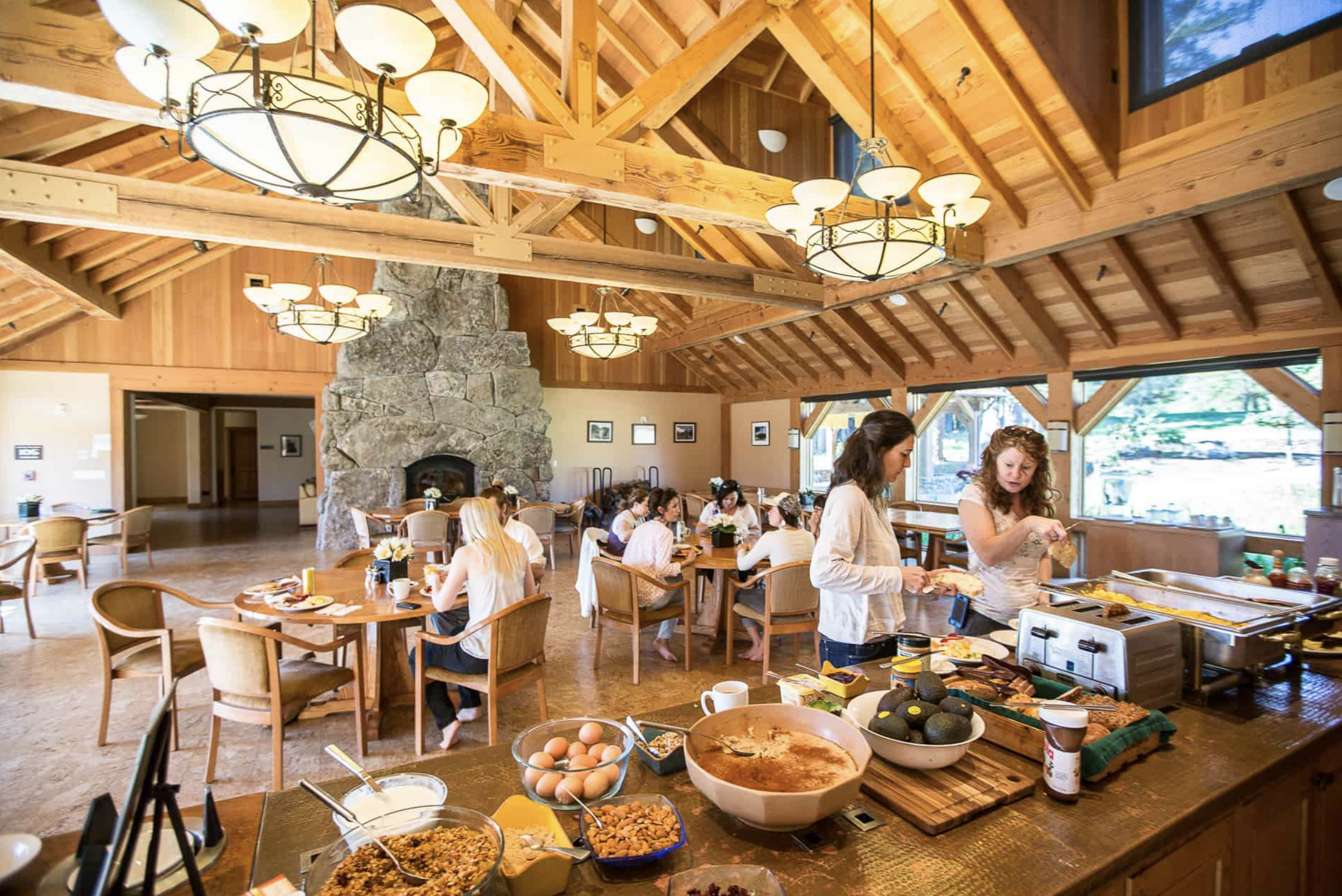 RatnaLing-TZ-5 A spacious dining area with wooden beams and a stone fireplace features people seated at tables enjoying a meal. Two women in the foreground prepare food at a buffet table with various breakfast items. The room has large windows and warm lighting.